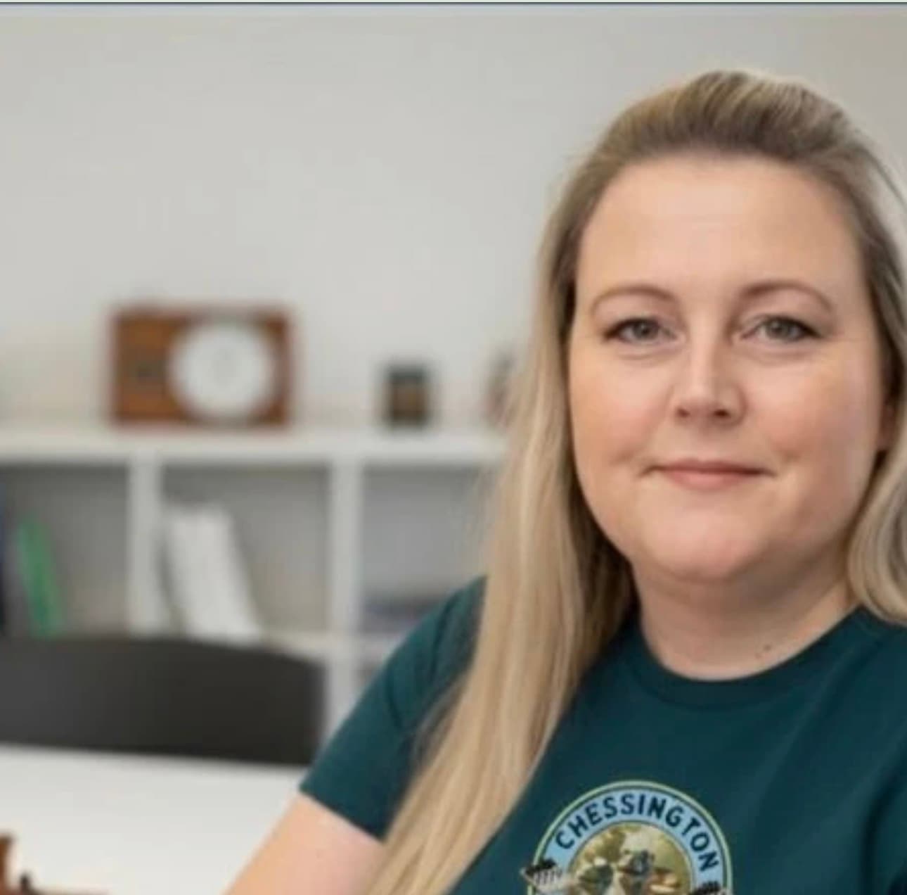 Smiling blonde woman wearing a green Chessington t-shirt in a blurred office setting.